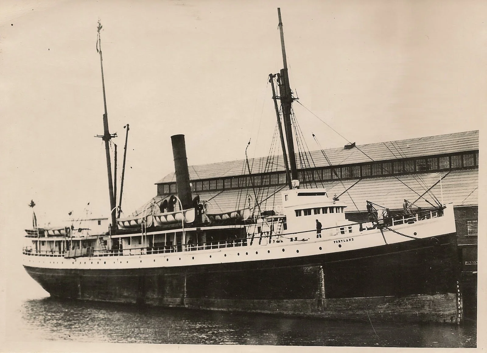 Vintage black and white photograph of steamship docked at waterfront warehouse with tall masts and smokestack